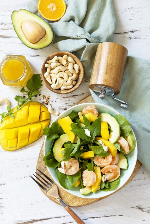 Vegetarian vegan healthy food. Salad with arugula, mango, avocado, shrimp, pecans and dressing of olive oil, honey and wine vinegar on a white rustic kitchen table. Top view, flat lay background.